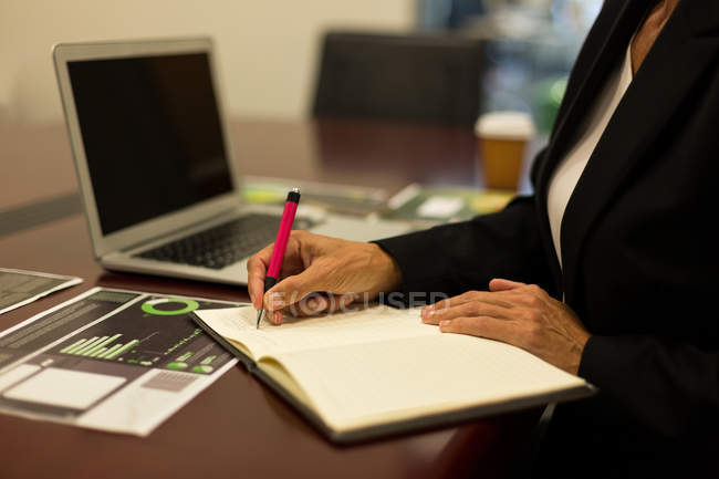 Sección media de la mujer de negocios madura escribiendo en diario en el escritorio en la oficina - foto de stock