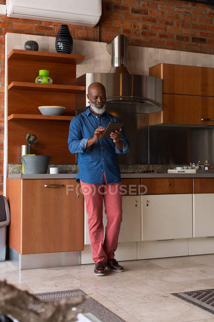 Старший мужчина, стоящий на кухне, используя цифровой планшет дома — стоковое фото