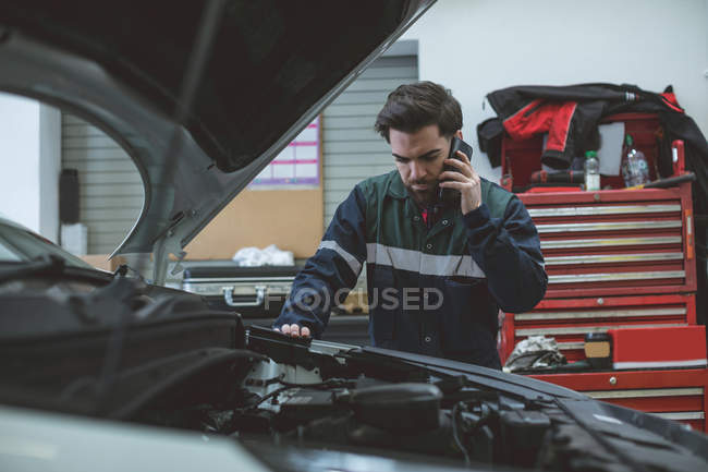 Mecánico hablando en un teléfono móvil mientras examina el coche en el garaje de reparación - foto de stock