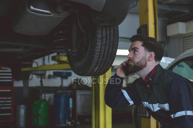 Mecânico masculino falando no telefone celular enquanto examina carro na garagem de reparação — Fotografia de Stock