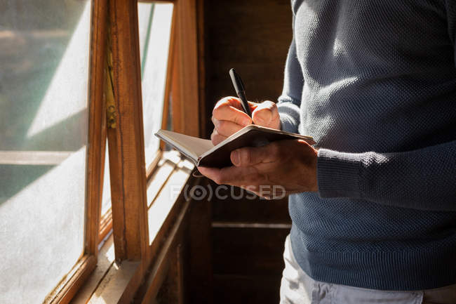 Sección media del hombre escribiendo nota en el diario en la cabaña de troncos - foto de stock