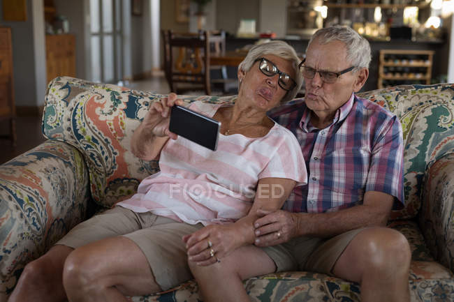 Pareja mayor tomando selfie con teléfono móvil en la sala de estar en casa - foto de stock
