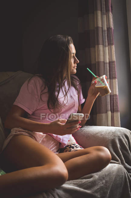Donna che prende il caffè freddo mentre usa il telefono cellulare a casa — Foto stock