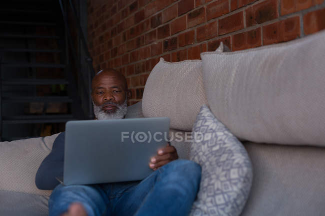 Hombre mayor acostado en el sofá mientras usa el ordenador portátil en casa - foto de stock