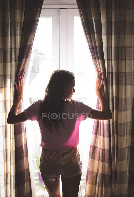 Vista posteriore della donna che apre le tende delle finestre a casa — Foto stock