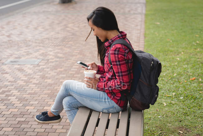 Mujer sonriente sentada en el banco y usando el teléfono móvil en el parque - foto de stock