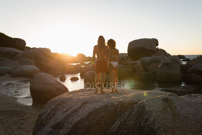 Vista trasera de la pareja de pie juntos en una roca en la playa - foto de stock