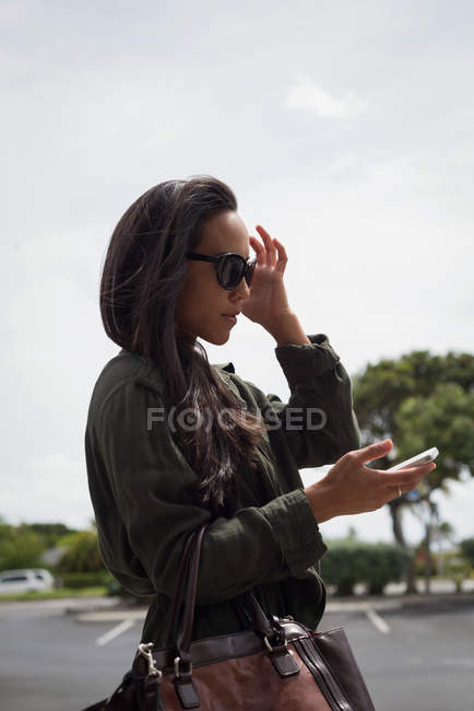 Красива жінка в сонцезахисних окулярах, використовуючи мобільний телефон на вулиці — стокове фото