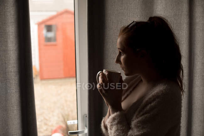 Задумчивая женщина пьет кофе, глядя в окно дома — стоковое фото