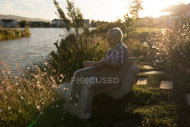 Старший мужчина отдыхает на скамейке у реки в солнечный день — стоковое фото