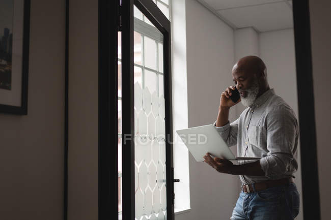 Senior graphic designer using laptop while talking on mobile phone in office — Stock Photo