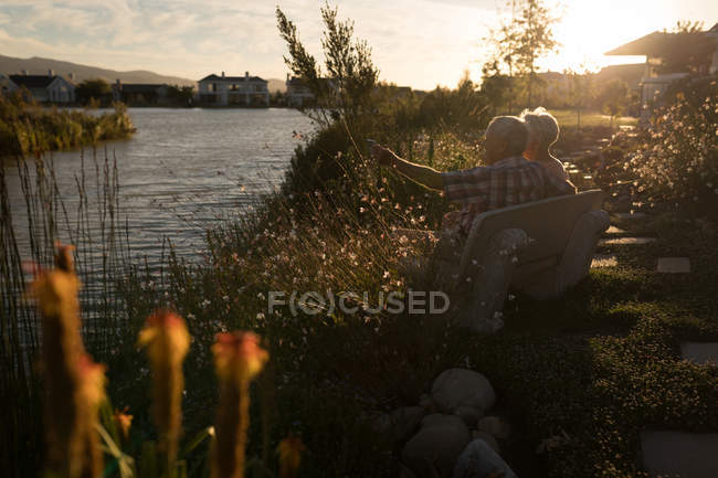 Pareja mayor relajándose en un banco cerca de la orilla del río en un día soleado - foto de stock