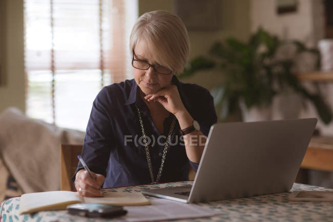 Зрелая женщина ведет дневник, используя ноутбук дома — стоковое фото