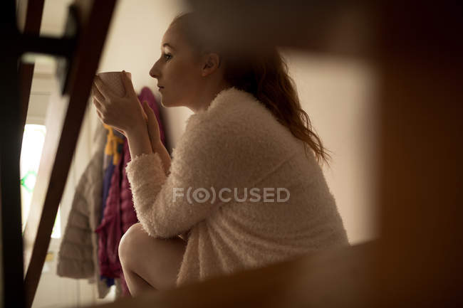 Mujer pensativa tomando café mientras se relaja en las escaleras en casa - foto de stock