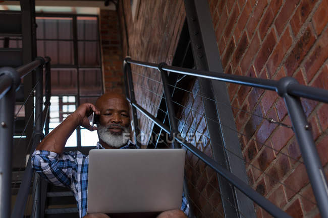 Homem sênior falando no telefone celular enquanto usa laptop na escada — Fotografia de Stock