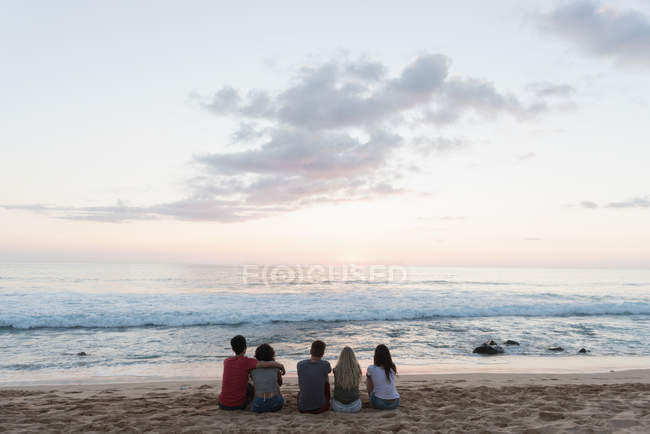 Grupo de amigos sentados juntos en la playa al atardecer - foto de stock
