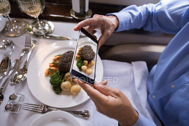 Sección media del hombre de negocios tomando fotos de la comida con teléfono móvil en jet privado - foto de stock