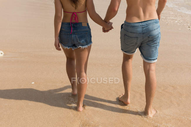 Bassa sezione di coppia che cammina insieme mano nella mano in spiaggia — Foto stock