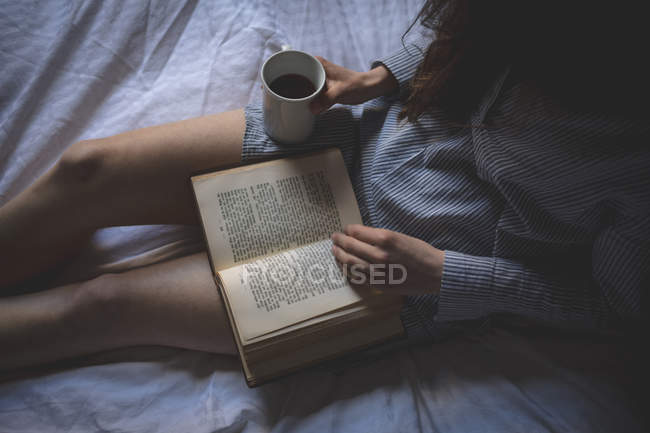 Woman with coffee reading a book on bed at home — Stock Photo