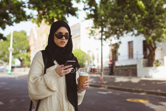 Hijab mujer usando el teléfono móvil mientras sostiene la taza de café en la calle - foto de stock