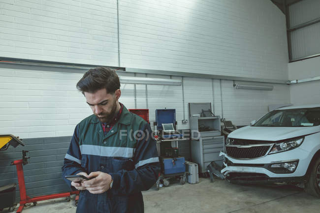 Mecánico utilizando el teléfono móvil durante la reparación de coches en el garaje - foto de stock