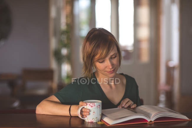 Красива жінка читає книгу, маючи каву вдома — стокове фото