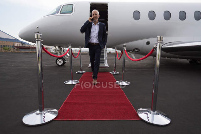 Empresario hablando por teléfono móvil mientras sale de jet privado en terminal - foto de stock