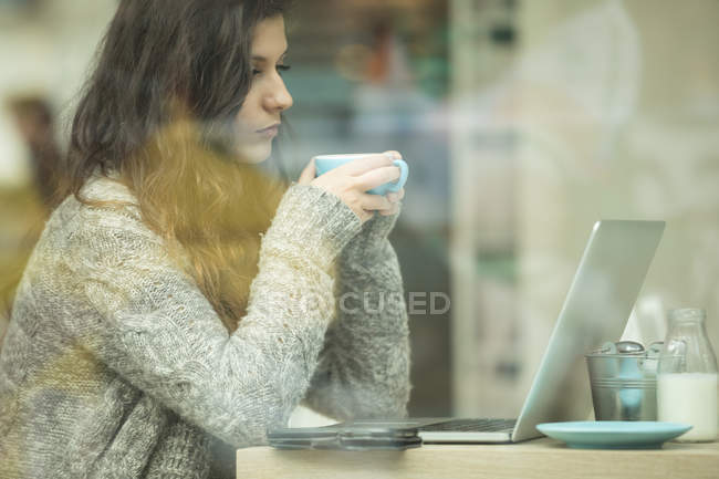 Молодая женщина пьет кофе во время использования ноутбука в кафе — стоковое фото
