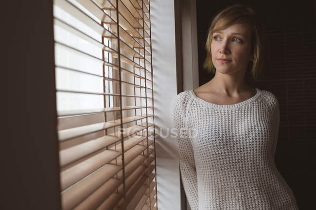 Bella donna che guarda attraverso la finestra cieca a casa — Foto stock