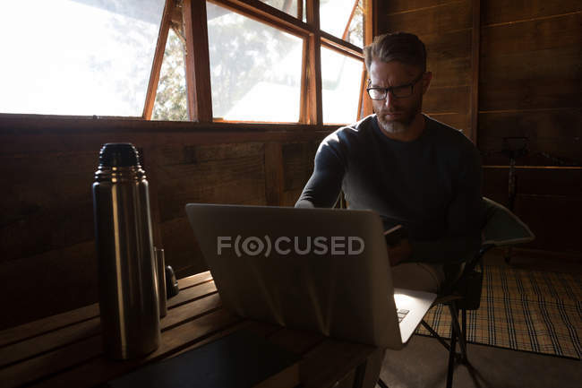 El hombre reflexivo usando el ordenador portátil en la cabaña de registro - foto de stock