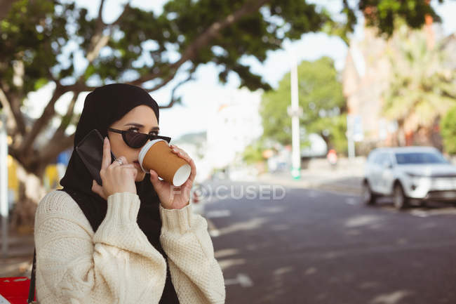 Hermosa mujer hijab hablando en el teléfono móvil mientras toma café en la cafetería del pavimento - foto de stock