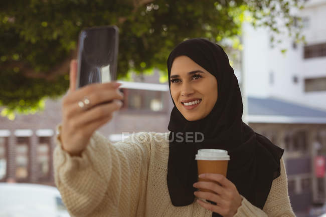 Усміхнена хіджаб жінка бере селфі з мобільним телефоном у тротуарному кафе — стокове фото