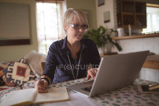 Зрілі жінки, використовуючи ноутбук під час запису в щоденнику вдома — стокове фото