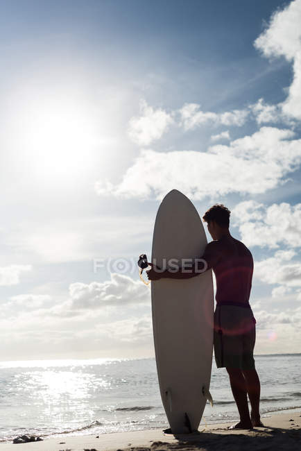 Vista trasera del surfista masculino de pie con tabla de surf en la playa - foto de stock