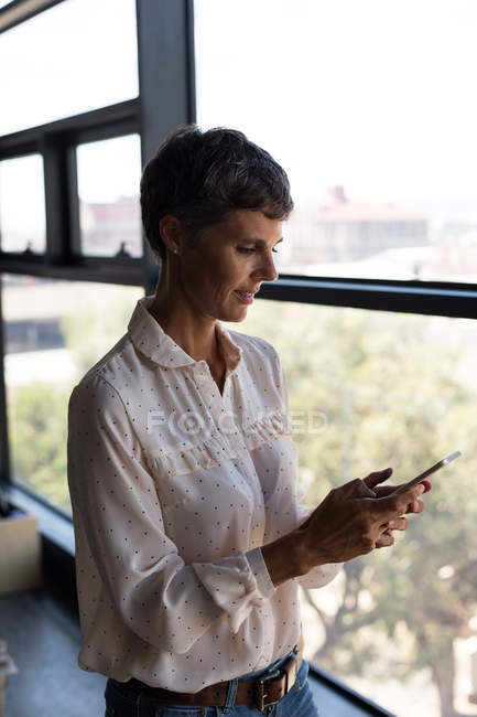 Улыбающаяся взрослая деловая женщина с мобильного телефона в офисе — стоковое фото