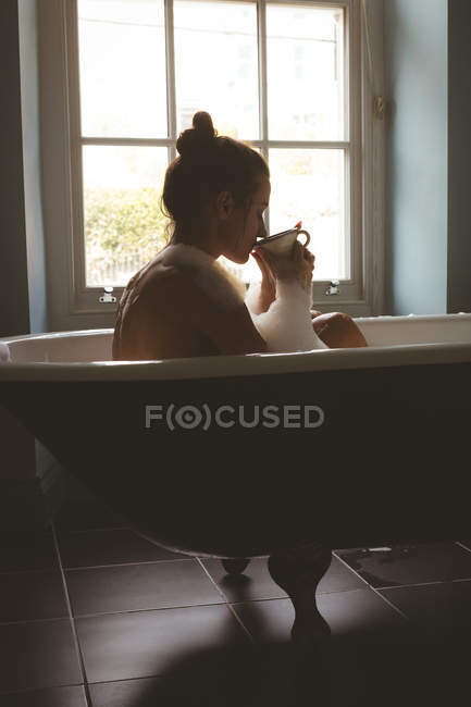 Mulher tomando café preto enquanto toma banho de espuma no banheiro — Fotografia de Stock