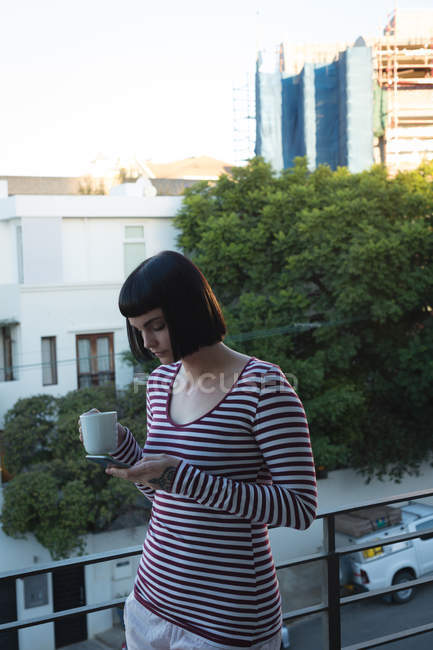 Жінка має каву під час використання мобільного телефону на балконі вдома — стокове фото