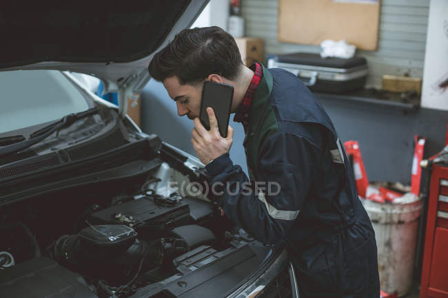 Mecânico falando em um telefone celular enquanto examina carro na garagem de reparação — Fotografia de Stock