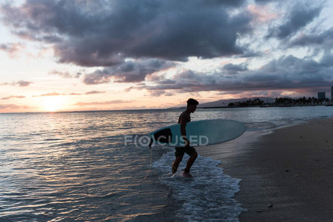 Hombre surfista de pie con tabla de surf en la playa al atardecer - foto de stock