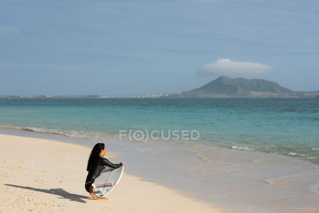 Femme accroupie avec planche de surf à la plage par une journée ensoleillée — Photo de stock