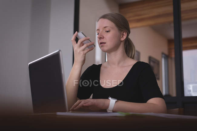 Female executive talking on mobile phone while working on laptop in office — Stock Photo