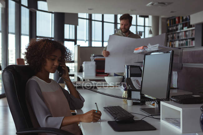 Женщина-архитектор разговаривает по телефону, пока коллега проверяет чертежи в офисе — стоковое фото
