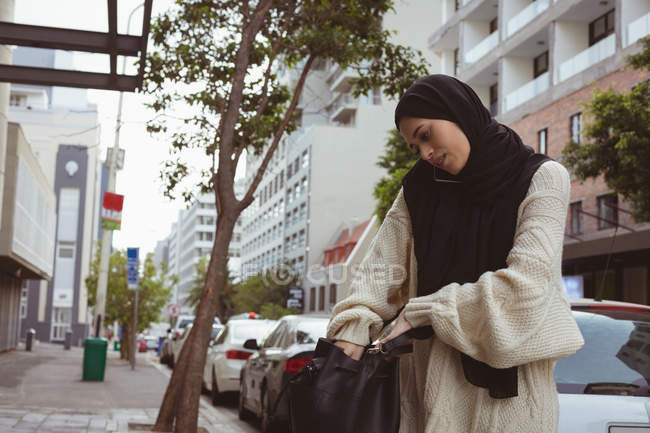 Hermosa mujer hijab hablando en el teléfono móvil mientras revisa su bolso - foto de stock
