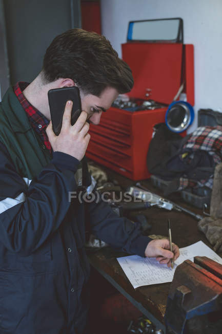 Mechaniker schreibt auf Papier, während er in der Werkstatt mit dem Handy telefoniert — Stockfoto