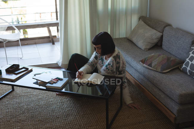 Mujer escribiendo en un libro en la sala de estar en casa - foto de stock