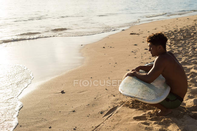 Surfista masculino sentado com prancha de surf na praia em um dia ensolarado — Fotografia de Stock