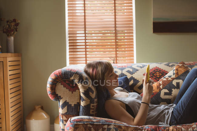 Mujer usando el teléfono móvil mientras está acostado en el sofá en la sala de estar - foto de stock