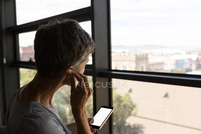 Mature businesswoman listening on headphones and looking through window at office — Stock Photo