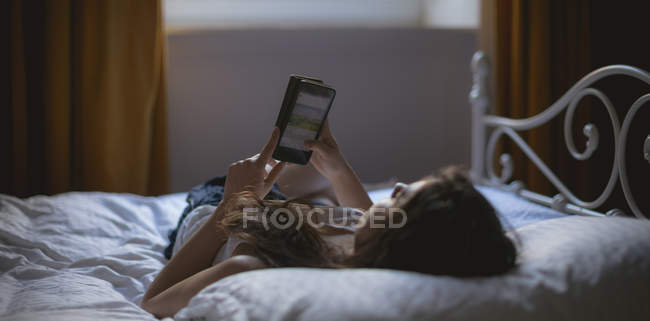 Frau benutzt Handy, während sie im Schlafzimmer auf dem Bett liegt — Stockfoto