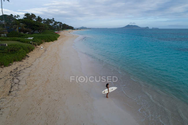 Mujer con tabla de surf de pie en la playa y mirando el mar - foto de stock